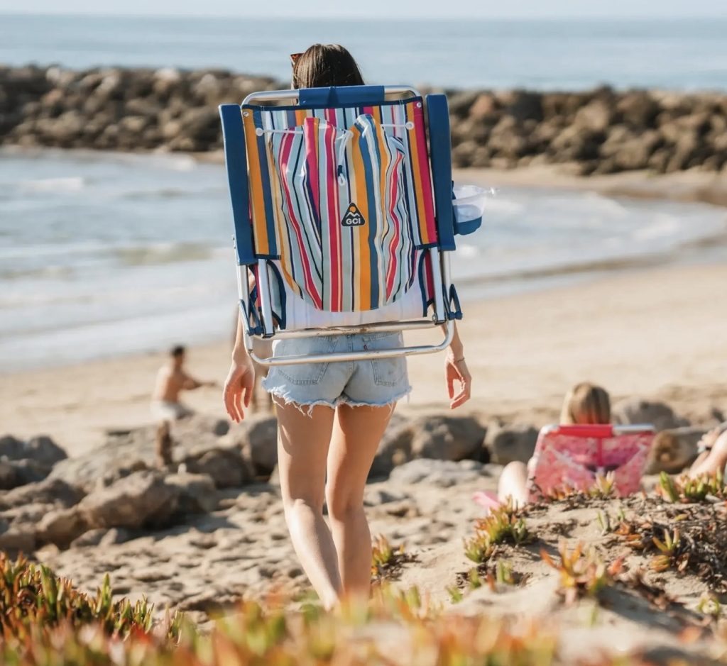 Person carrying a colorful striped GCI Outdoor Backpack Beach Chair on their back, walking toward the ocean on a sandy beach